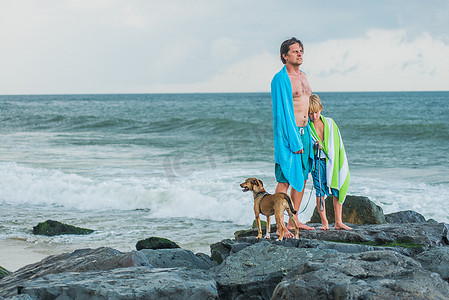 父子俩站在海边的岩石上身上披着沙滩毛巾旁边是宠物狗