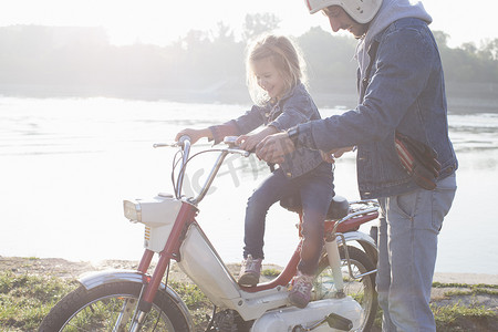 职场女孩摄影照片_年轻女孩坐在父亲的轻便摩托车上父亲站在她身边湖边