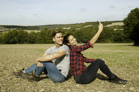 年轻的徒步旅行夫妇背靠背坐在田野里自拍智能手机英国白金汉郡大米森登