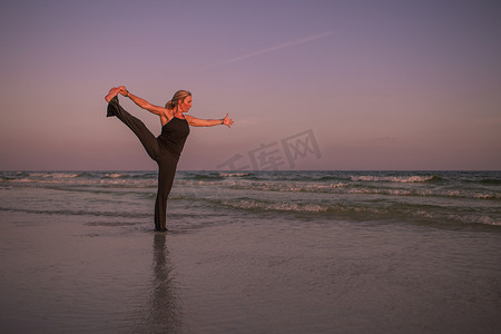 运动裤女摄影照片_日落时在海滩上练习瑜伽姿势的成熟女性