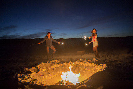 两个年轻女孩在篝火旁的海滩上拿着火花拿着火花