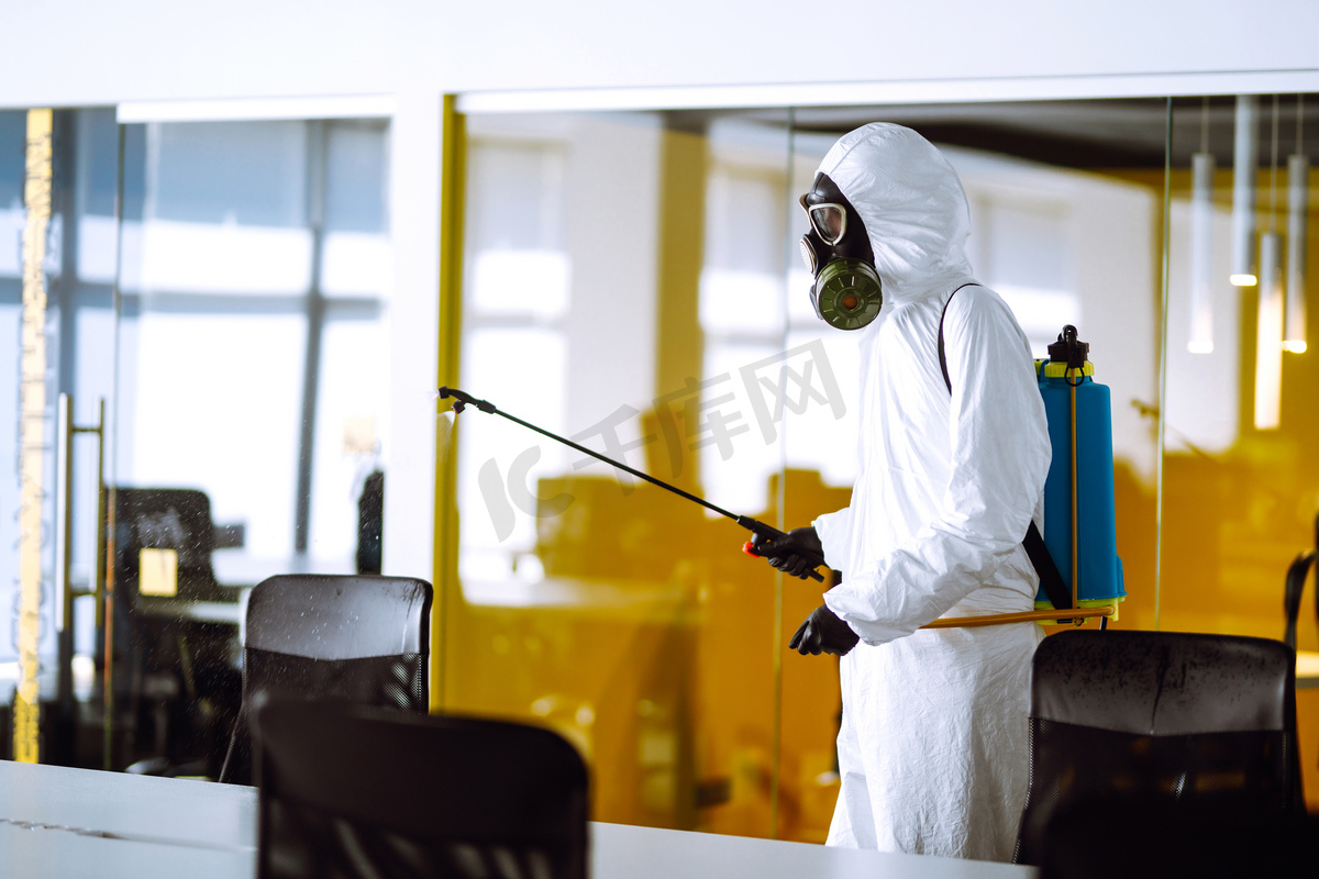 办公室消毒以防止COVID-19，穿着防护服的男子用喷雾剂化学品，以防止大肠病毒在检疫城市的蔓延。清洗概念.图片