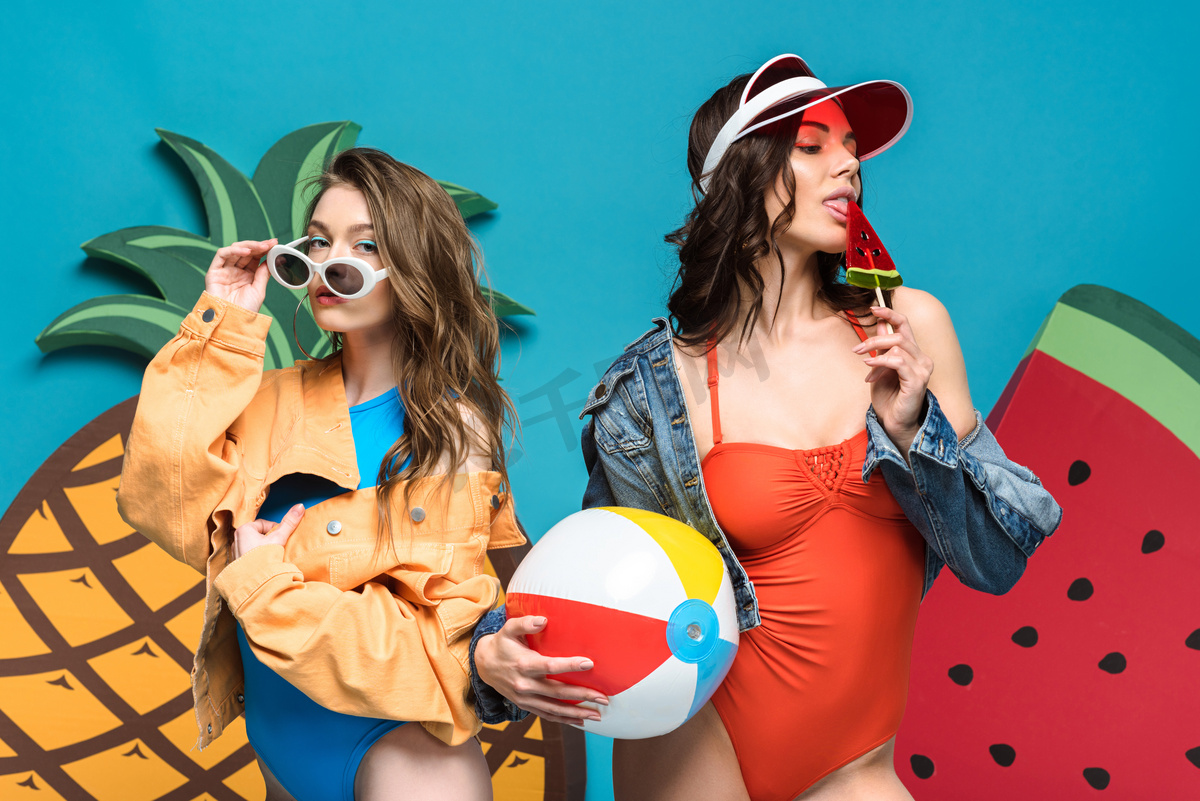 两个女孩与沙滩球和棒棒糖附近的装饰西瓜和菠萝隔离在蓝色图片