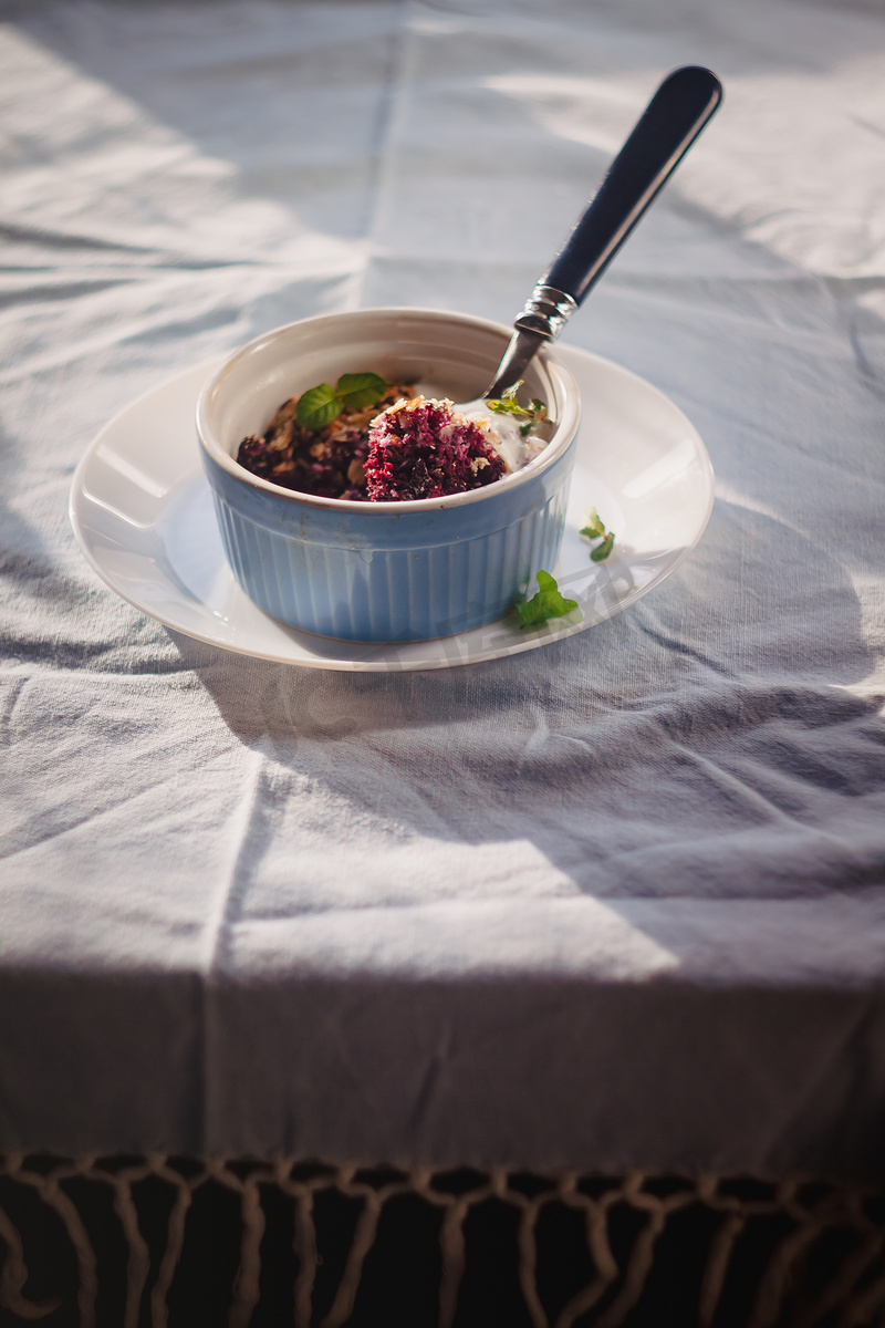 质朴的亚麻桌布上的白色和蓝色盘子里放着薄荷烤燕麦蓝莓碎屑。图片