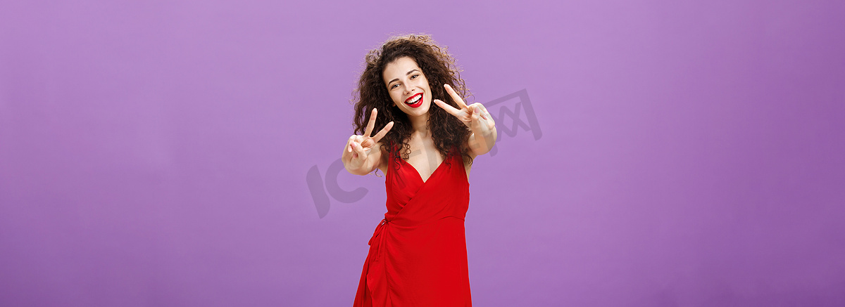 长相友好和平的欧洲女性，卷发，穿着优雅的红色连衣裙，展示着和平或胜利的迹象，大笑着说她在紫色墙壁上的朋友圈里享受很棒的派对图片
