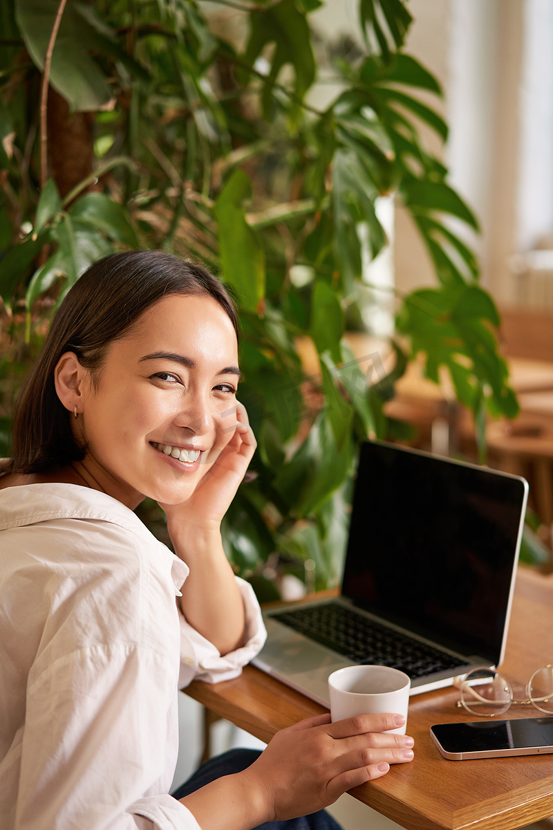 时髦的年轻女性坐在咖啡馆里，对着镜头微笑、喝咖啡、使用笔记本电脑、远程工作、学习或浏览互联网图片