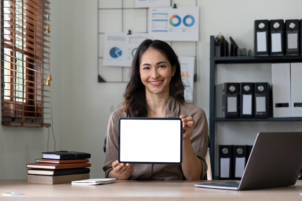 快乐的亚洲女人坐在办公室时展示带有空白屏幕的平板电脑，为广告制作模型。图片