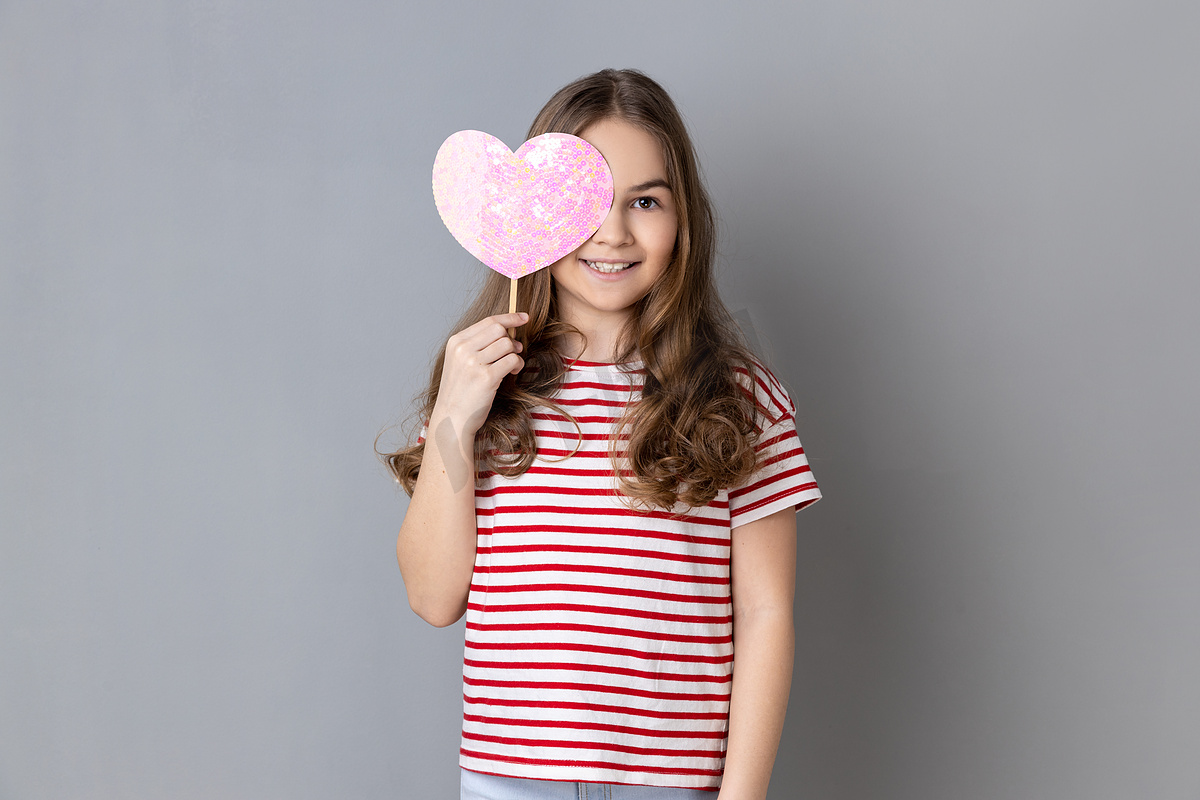 穿着条纹T恤的小女孩用棍子上的粉色心遮住眼睛，表现出浪漫的情怀。图片