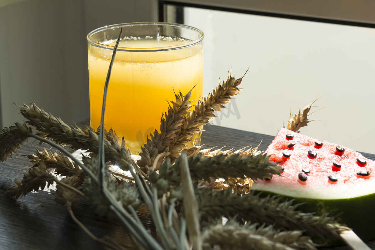 阳光下的木桌上放着鲜榨橙汁、甜西瓜甜点和成熟小麦的小穗。图片