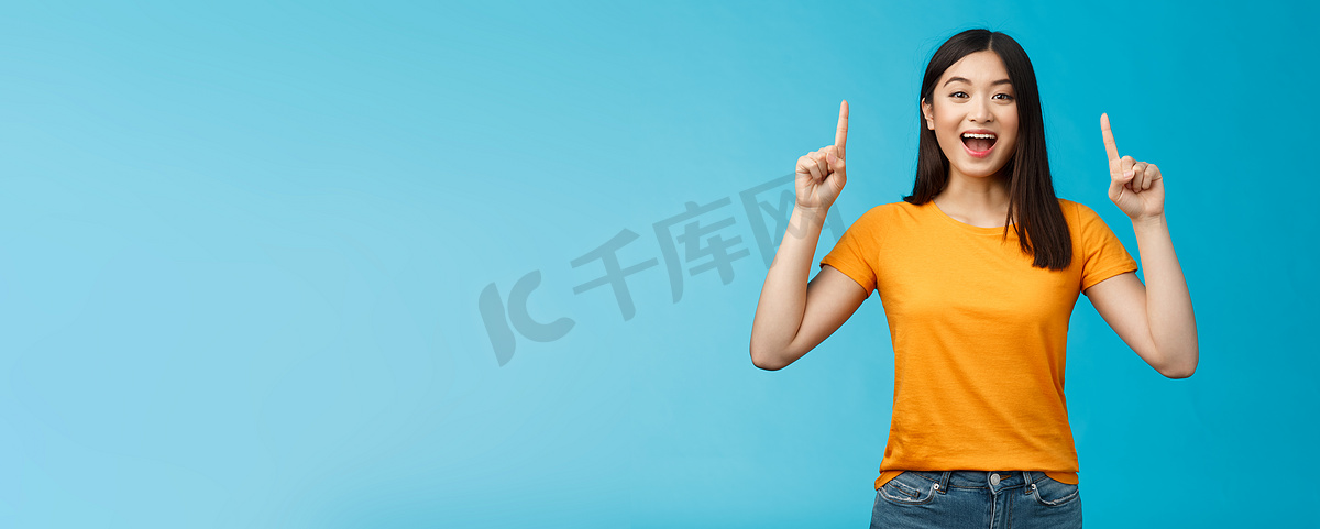 有趣的亚洲女孩向你介绍新产品分享促销，举起双手指指点点地微笑，看起来兴奋和乐观，热情的广告，站在蓝色背景图片
