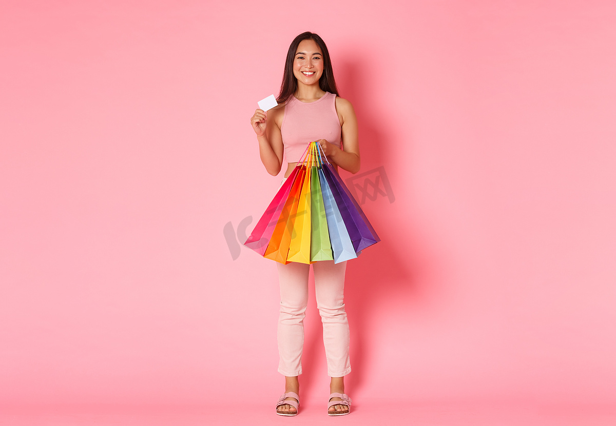 快乐时尚的亚洲女孩的全长肖像展示她的折扣信用卡和拿着购物袋，享受花钱买新衣服，站在粉红色的背景上图片