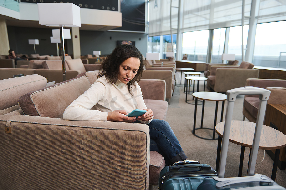 漂亮女人使用手机，浏览网站，预​​订酒店，同时在贵宾休息室舒适的沙发上休息，在机场等待航班图片