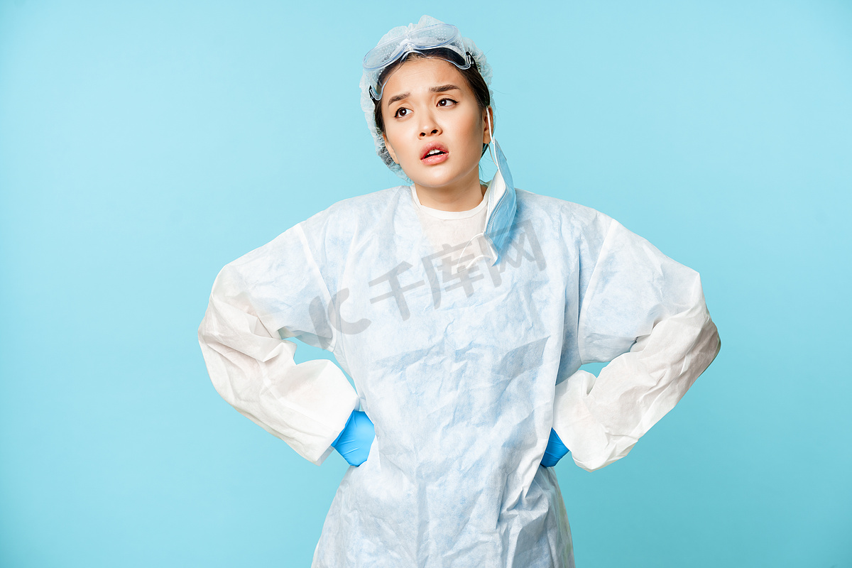 疲惫的亚洲护士或医生摘下医用口罩，感到疲劳，穿着个人防护装备，双手叉腰叹息，蓝色背景图片