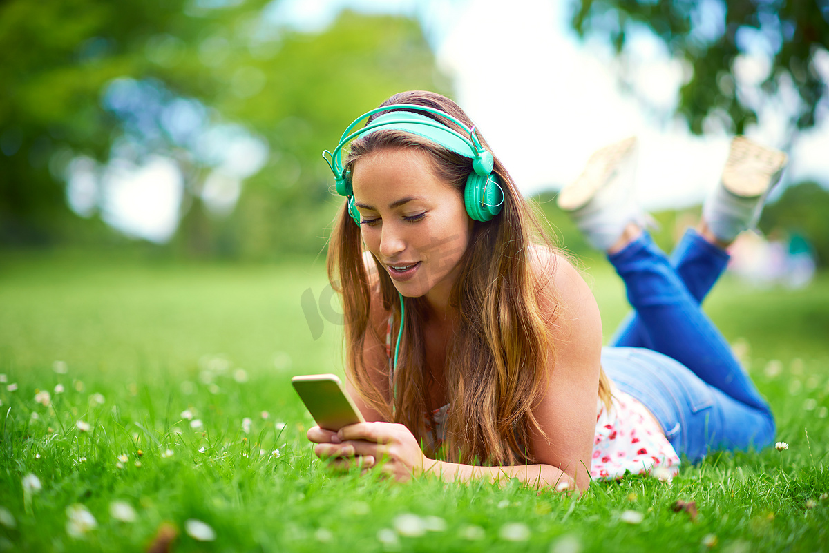 音乐是最甜蜜的逃避……一位年轻女子躺在公园的草地上听音乐。图片