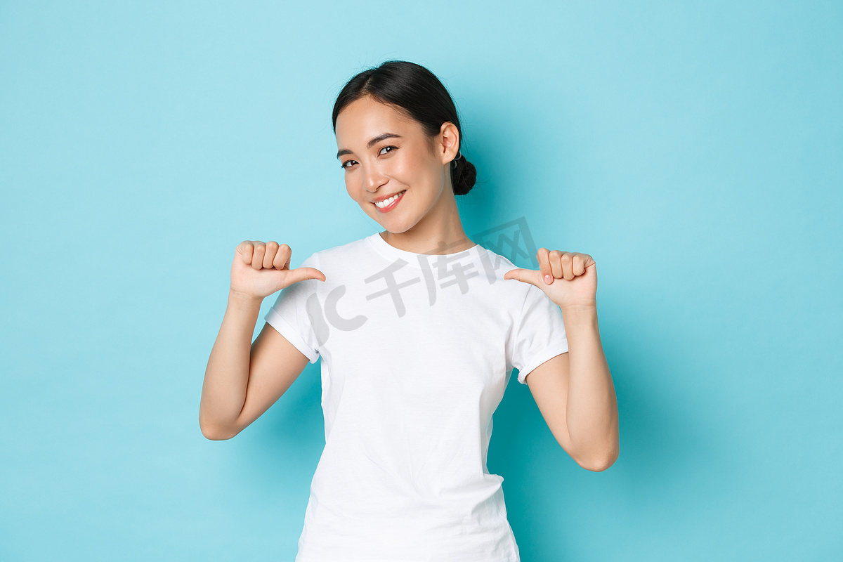 身穿白色T恤、自信微笑的亚洲女性用自豪、自信的表情指着自己，炫耀，提升自己的能力，寻找工作，站在蓝色背景图片