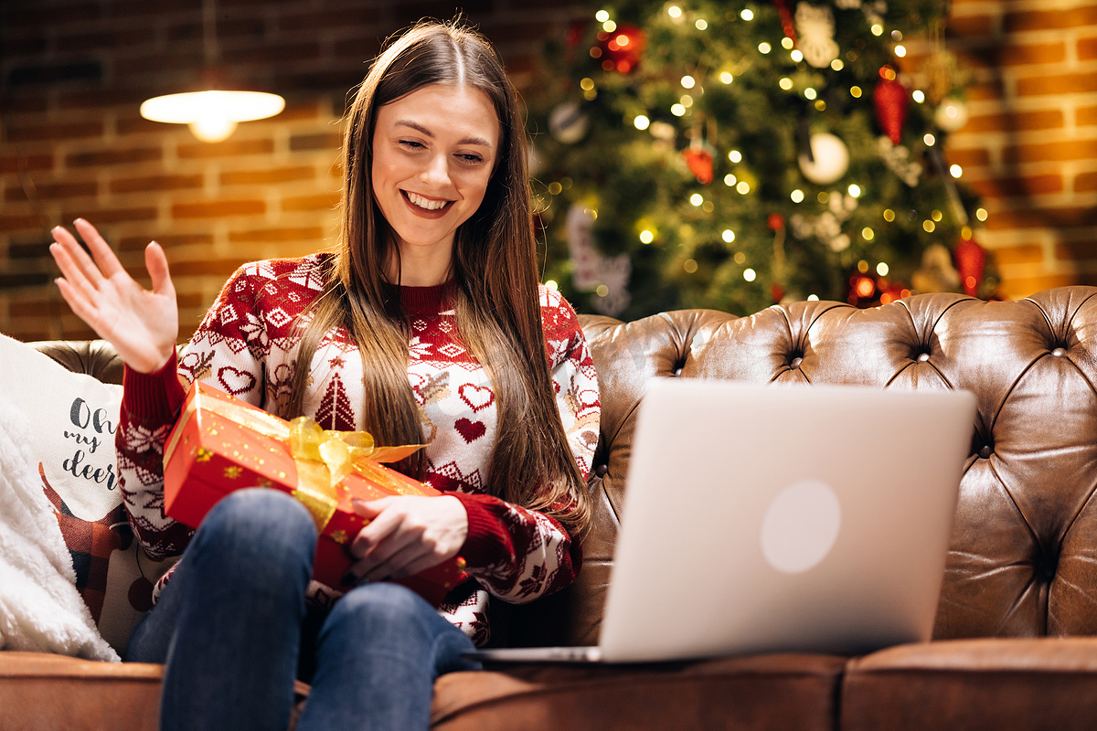 兴奋快乐的白人女孩戴着圣诞老人的帽子拿着圣诞礼盒，在电脑视频通话虚拟会议聊天中打开圣诞礼物与家人聊天，社交距离朋友坐在家里的沙发上图片