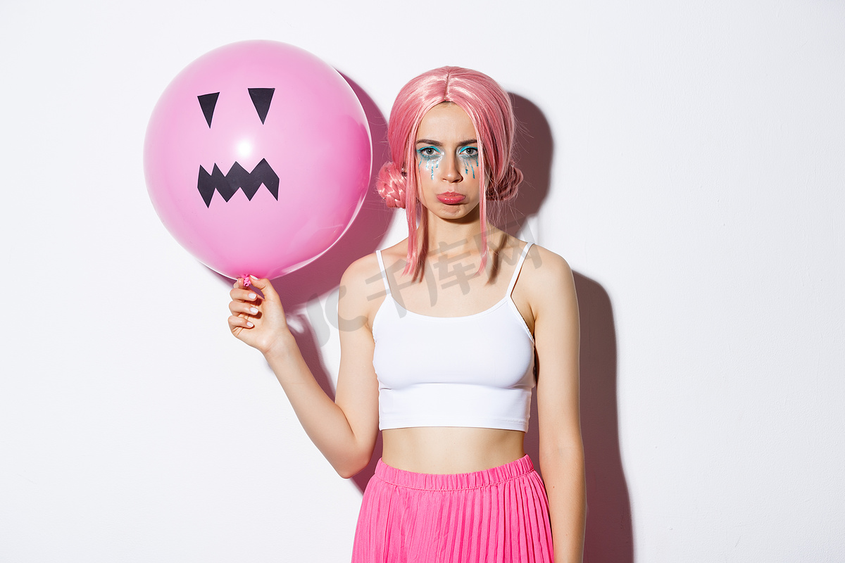 一张阴郁失望的派对女孩的照片，她化着鲜艳的妆容，戴着粉色假发，拿着杰克灯笼脸的气球，庆祝万圣节图片