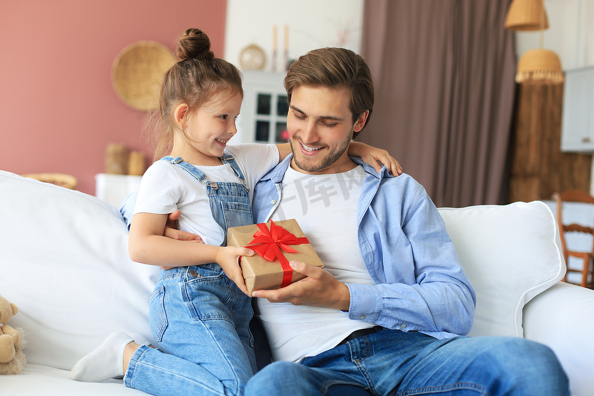 可爱的小女儿给爸爸惊喜，小女孩把礼盒送给坐在沙发上的爸爸。图片