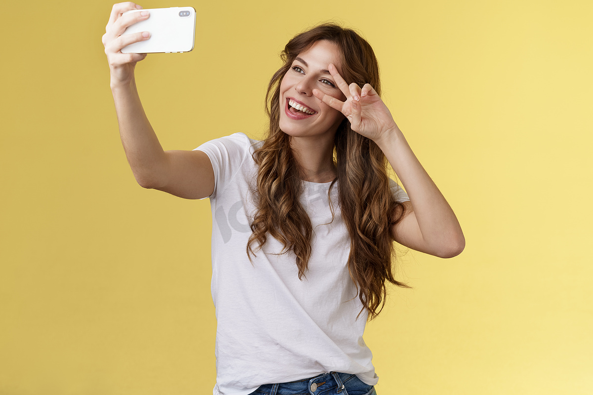 欢快的魅力帅气的卷发女性白色 T 恤展示和平胜利的标志，微笑着广泛摆出温柔友好的表情智能手机前置摄像头自拍黄色背景图片
