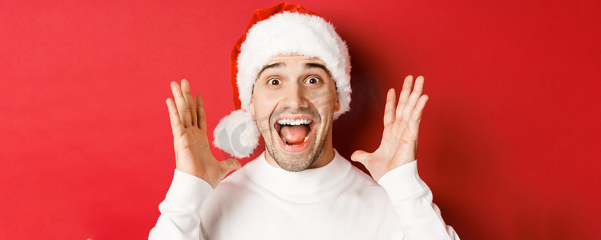 戴着圣诞帽的快乐年轻人的特写，发布重大的圣诞公告，微笑着惊讶，站在红色背景上图片