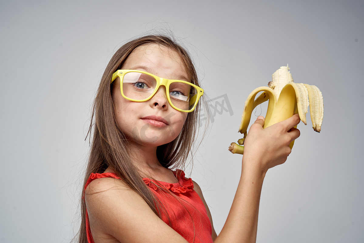 戴眼镜的小女孩，穿着红色连衣裙，手里拿着香蕉，浅色背景图片