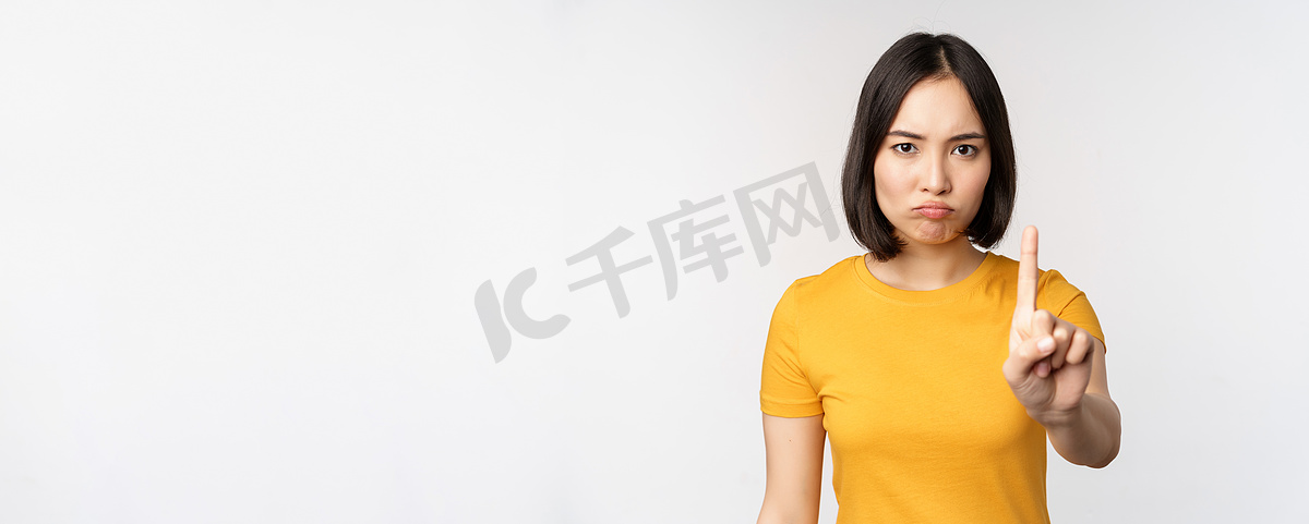 亚洲女性的肖像看起来严肃而愤怒，展示停止禁止手势、禁忌标志、禁止某物、站在白色背景上的黄色 T 恤图片