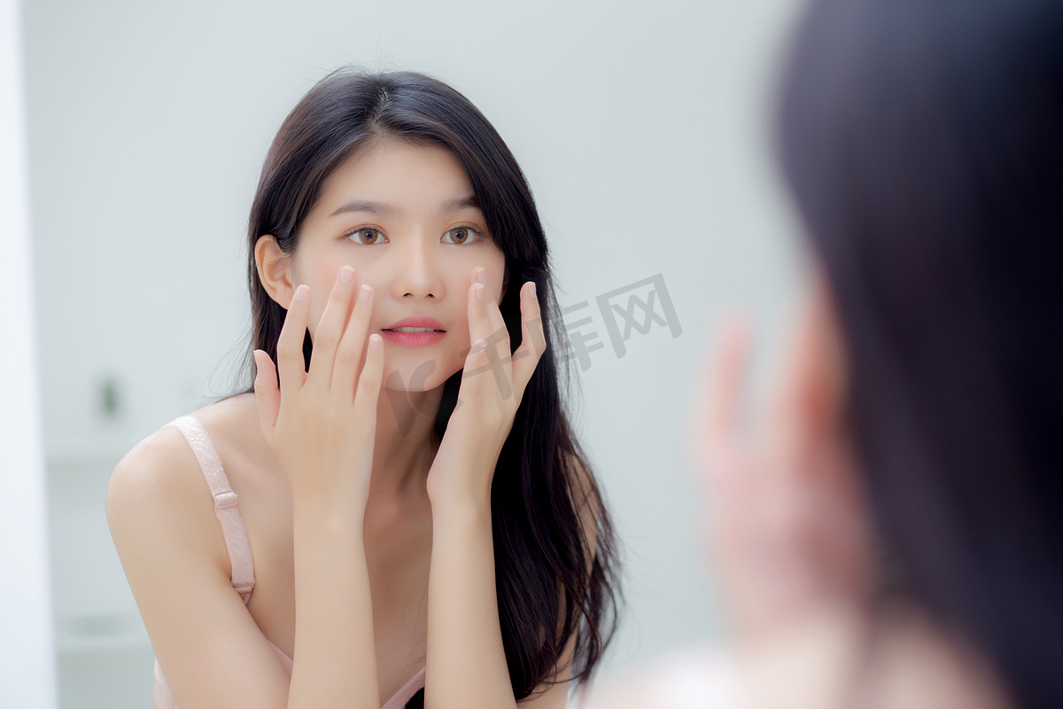 美丽的年轻亚洲女性微笑着看着镜子，用皮肤护理和化妆品检查脸部，以恢复活力和卫生，美丽的女孩快乐地用面霜或乳液清洁面部，以保持健康。图片