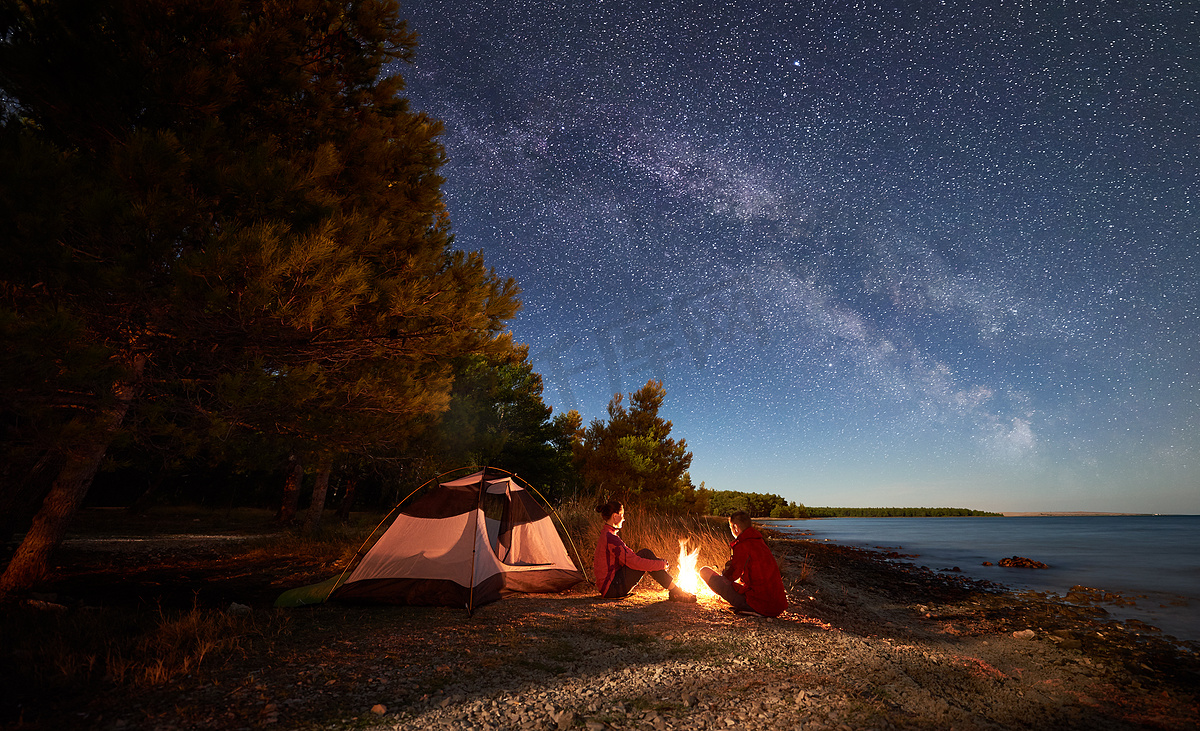 晚上在岸上露营。男子和女子徒步旅行者在帐篷前休息, 在黄昏的天空中充满了星星和银河的蓝色水和森林背景下的篝火。户外生活方式概念图片