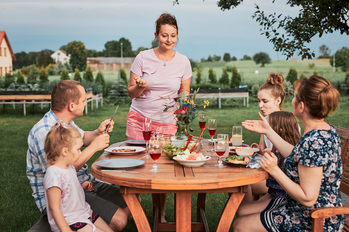 夏天外出野餐时,一家人在自家花园的烤架边吃饭.围坐在餐桌旁，端着食物和碗碟的人图片