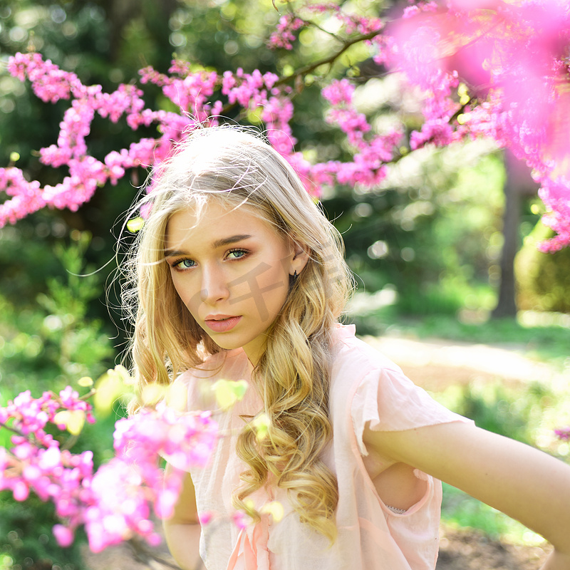 女孩在梦幻般的脸上, 温柔的金发碧眼紫花的犹大树, 自然背景。春天绽放概念。在阳光明媚的春天, 女士在公园散步。弥散的年轻女子在花园里欣赏花朵。.图片