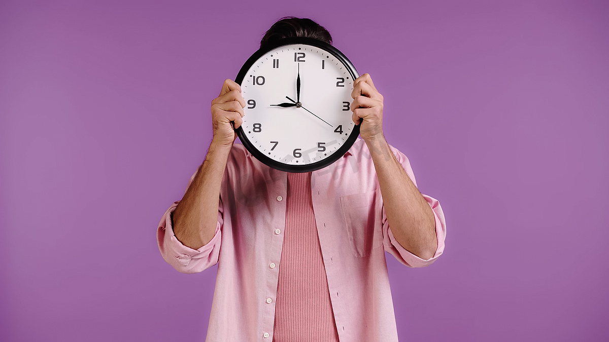 young man in pink shirt obscuring face with clock isolated on purple图片