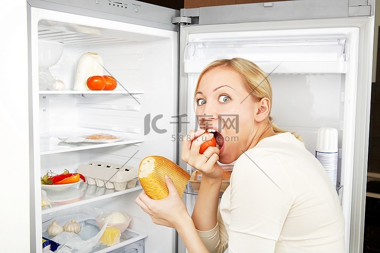 贪婪的女人靠着开着的冰箱吃饭
