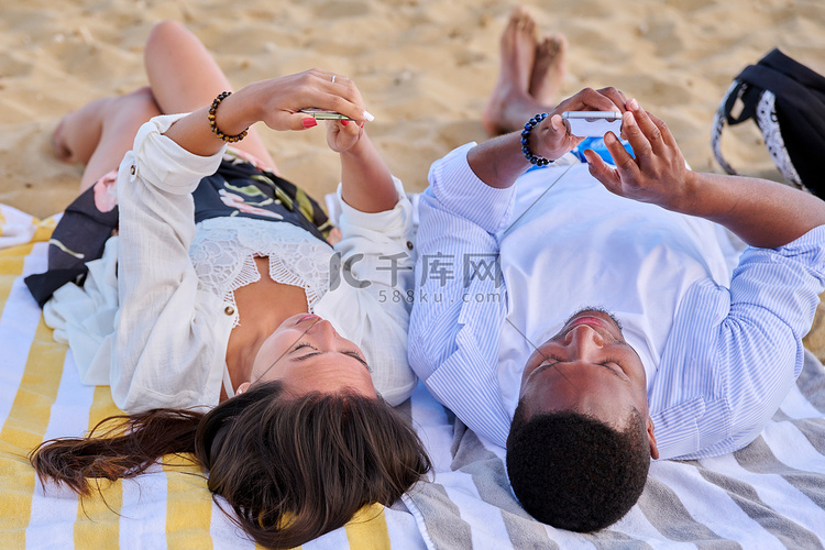 年轻的多文化夫妇躺在沙滩上使用