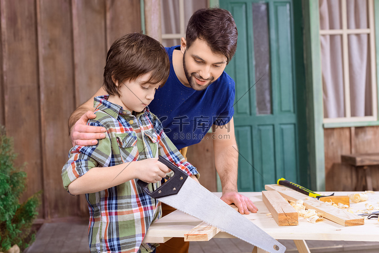 微笑的父亲看着专注的儿子锯木板