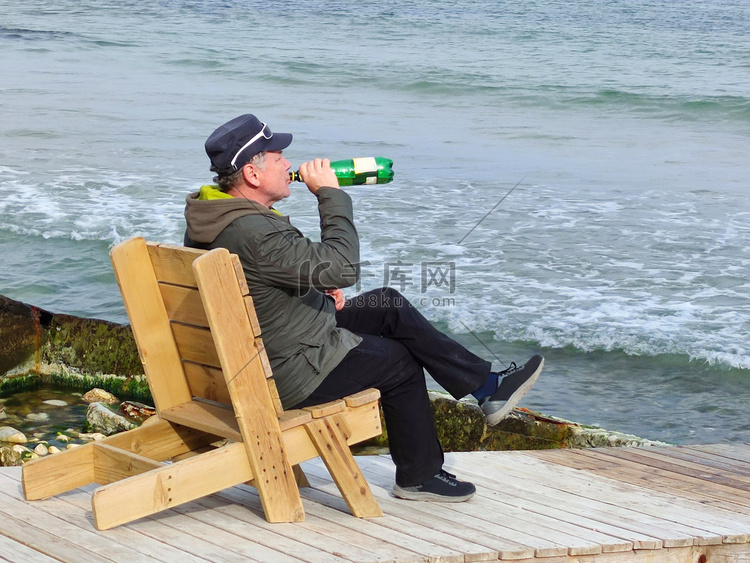 一个男人坐在海边的木制躺椅上喝