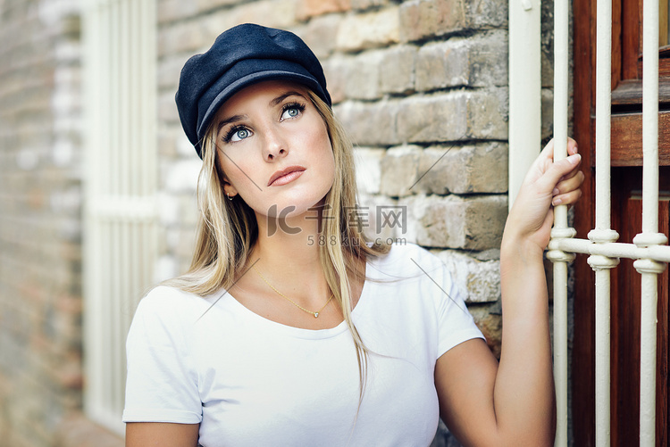 年轻的金发女人戴着帽子站在砖墙