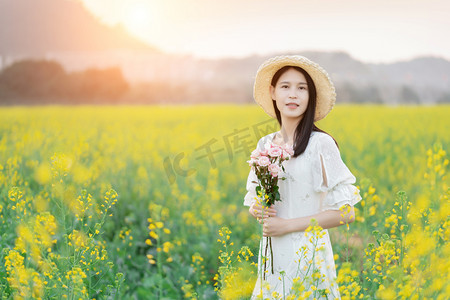 金黄色田野摄影照片_户外阳光下油菜花海中手捧鲜花的美丽女子摄影图配图