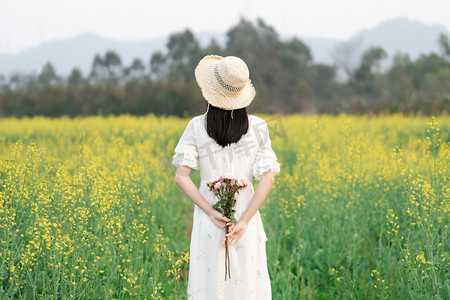 户外油菜花海中手捧鲜花少女的背影摄影图配图