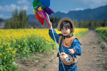 古代小孩坐桥边摄影照片_乡村春天女孩风车帽子田边玩风车摄影图配图