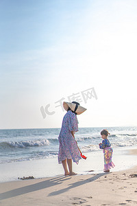 初春换新摄影照片_海边妈妈和小美人鱼下午母亲和女儿沙滩看海侧面摄影图配图