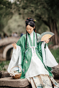 春游美女摄影照片_汉服美女白天汉服美女户外公园拿着扇子低头摄影图配图