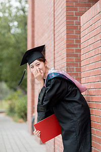 大学毕业照摄影照片_毕业季白天穿着学士服的美女教学楼捂嘴巴摄影图配图