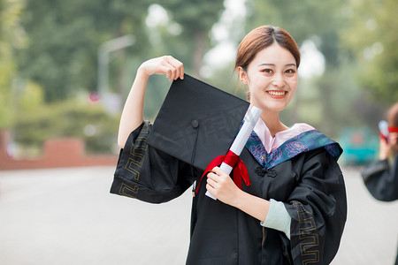 毕业季白天穿着学士服的美女户外拿起学士帽摄影图配图