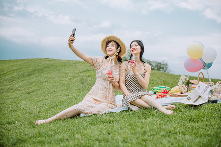 闺蜜野餐摄影照片_野餐郊游白天端着酒杯的闺蜜俩户外草地拿起手机自拍摄影图配图