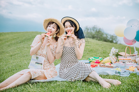 春游的摄影照片_野餐郊游白天坐在草地上的两位美女草地吃西瓜摄影图配图