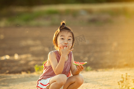 夏日暑假摄影照片_夏日儿童人像夕阳可爱小男孩田地旁蹲着拿西瓜吃手摄影图配图