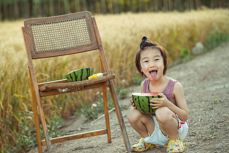 活泼孩子摄影照片_夏日儿童人像白天长发小男孩麦田抱西瓜调皮吐舌头摄影图配图