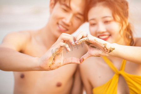 海边沙滩度蜜月摄影照片_爱情蜜月白天一对青年情侣沙滩比心摄影图配图
