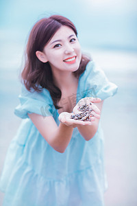 夏天美女白天穿着蓝色连衣裙的美女户外沙滩捧着贝壳摄影图配图