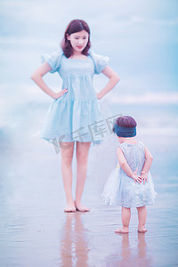 蓝色沙滩摄影照片_亲子白天模仿妈妈动作的小女孩沙滩面对面摄影图配图